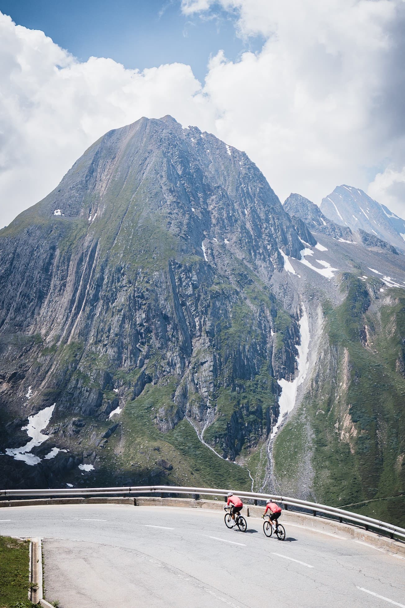 Two cyclists on an alpine road below a rocky peak